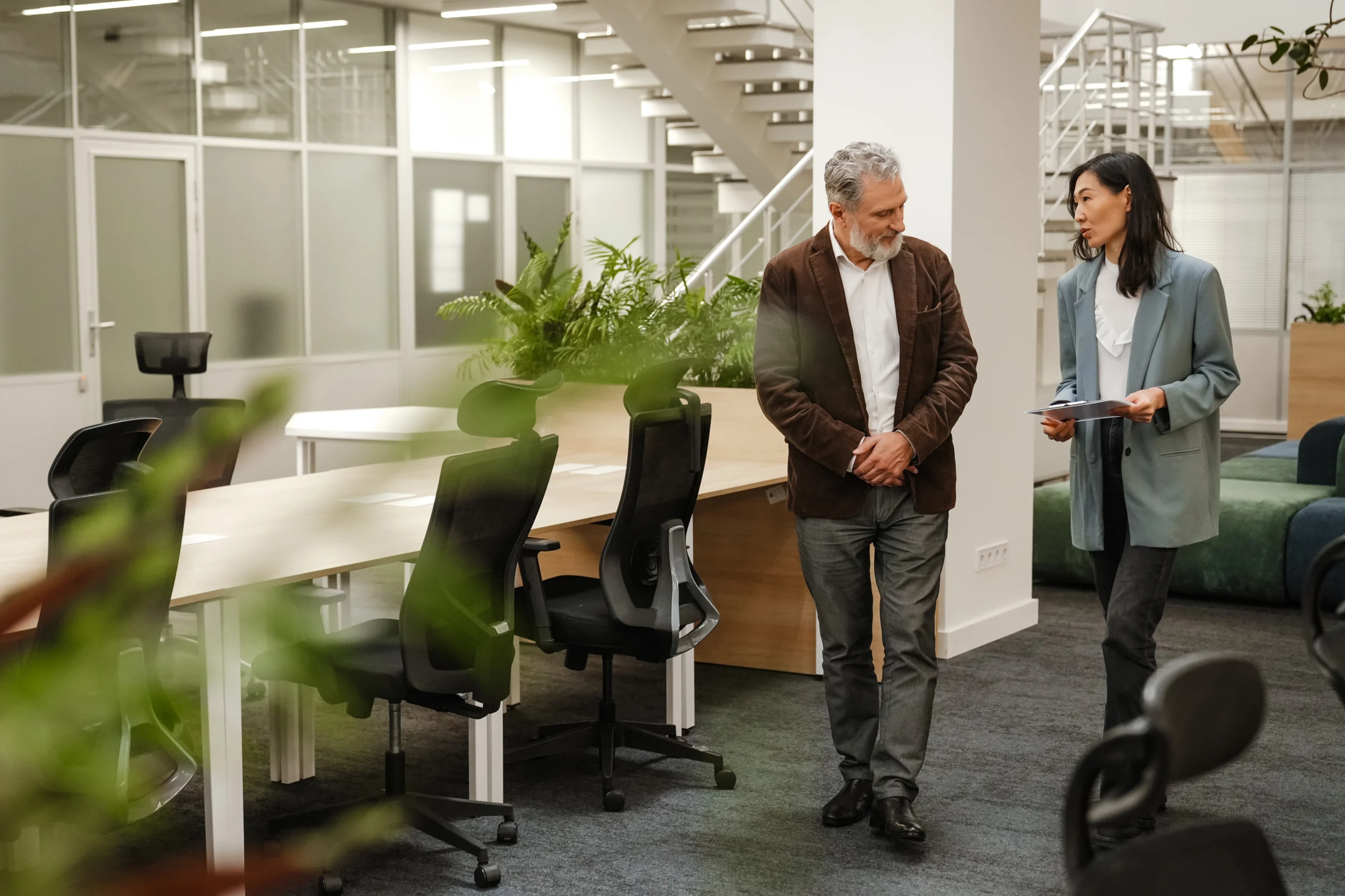 A middle-aged man and woman are walking and discussing documents in a modern open-plan office. They are business partners engaged in a professional conversation near workstations.