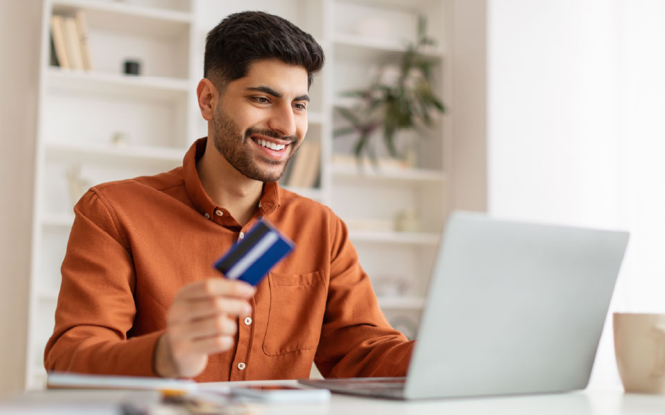 A smiling man in an orange shirt holds a blue credit card while looking at a laptop screen, ready to make an online purchase.